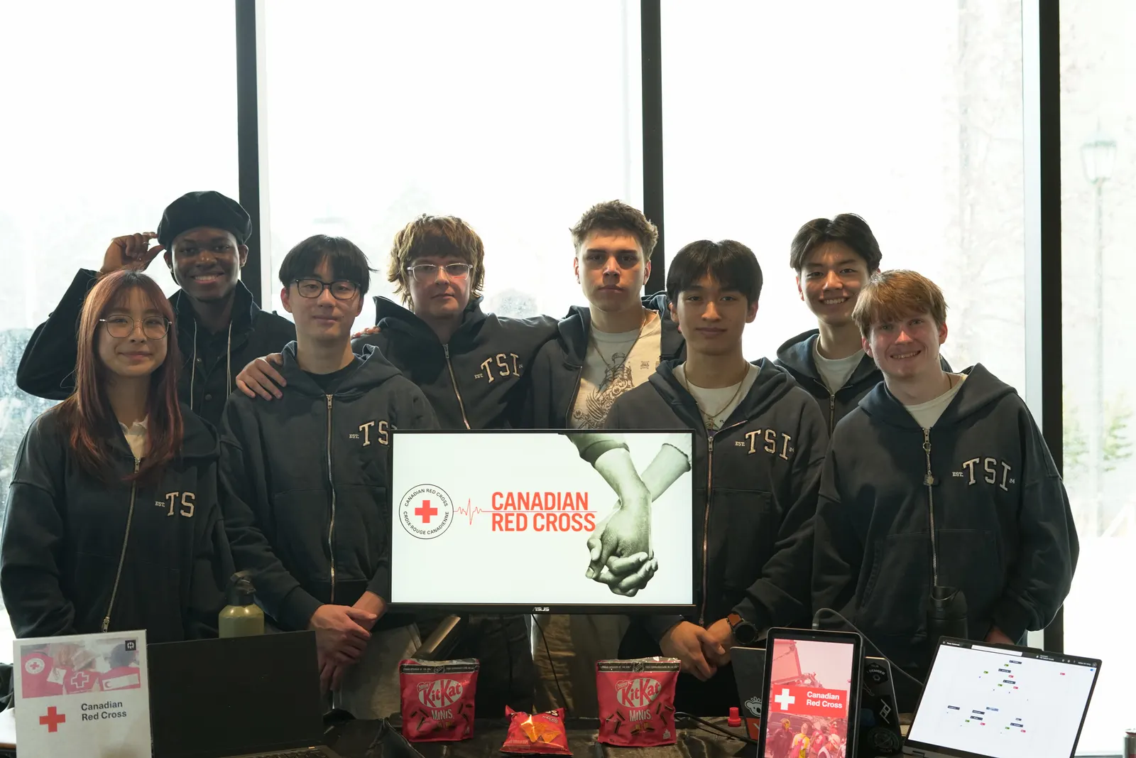 The eight-person Canadian Red Cross dev team in matching TSI hoodies behind their demo day booth, with a Canadian Red Cross title slide on the monitor