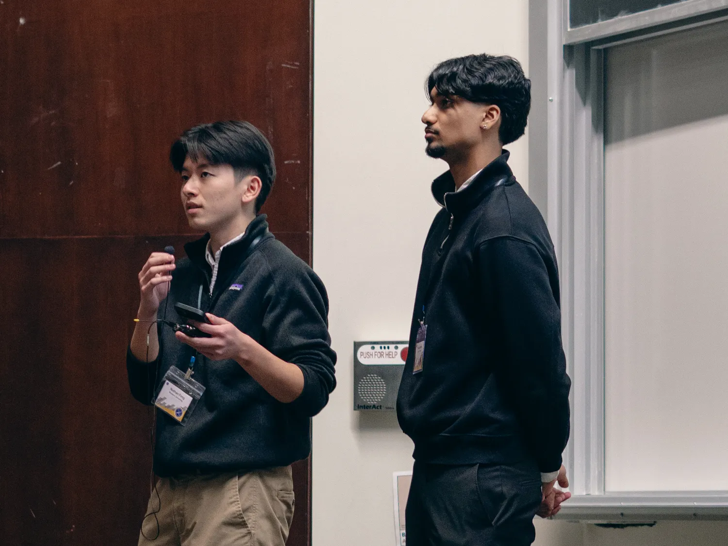 Nathan speaking into a microphone and holding a slide clicker at the front of a lecture hall, with a teammate standing beside him
